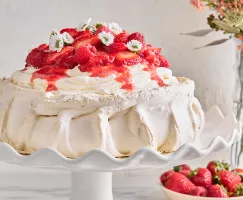 Pavlova topped with raspberries and strawberries and flowers on a white cake stand, shown with a vase of flowers and a bowl of berries