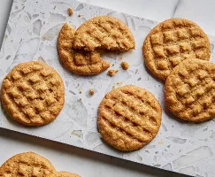 Seven golden-brown peanut butter cookies with distinctive fork press patterns displayed on parchment paper over a marble surface, one partially eaten to reveal a soft, chewy texture.