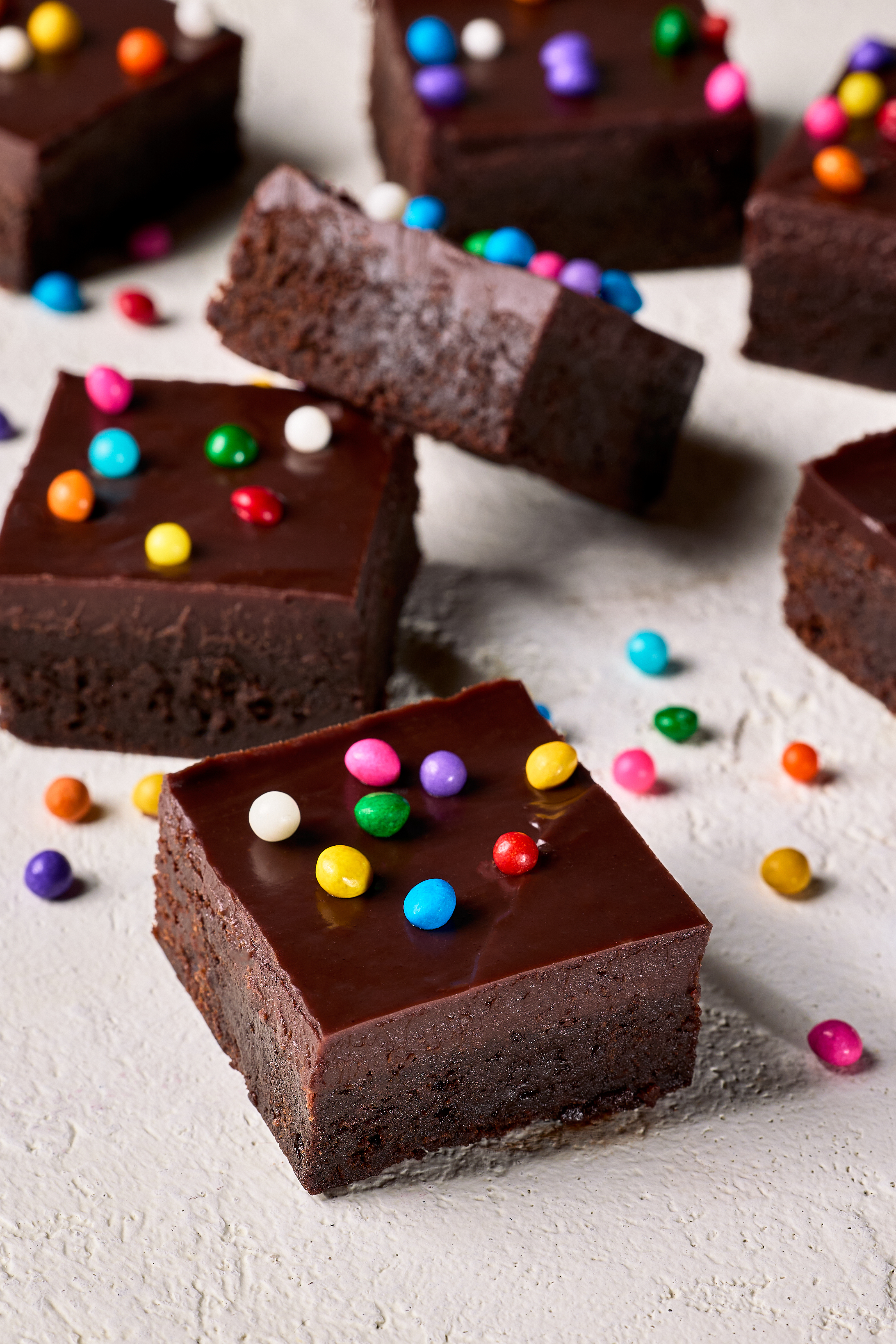 Square-cut Cosmic-style chocolate brownies randomly spread on waxed paper, topped with chocolate ganache and coloured candy sprinkles.