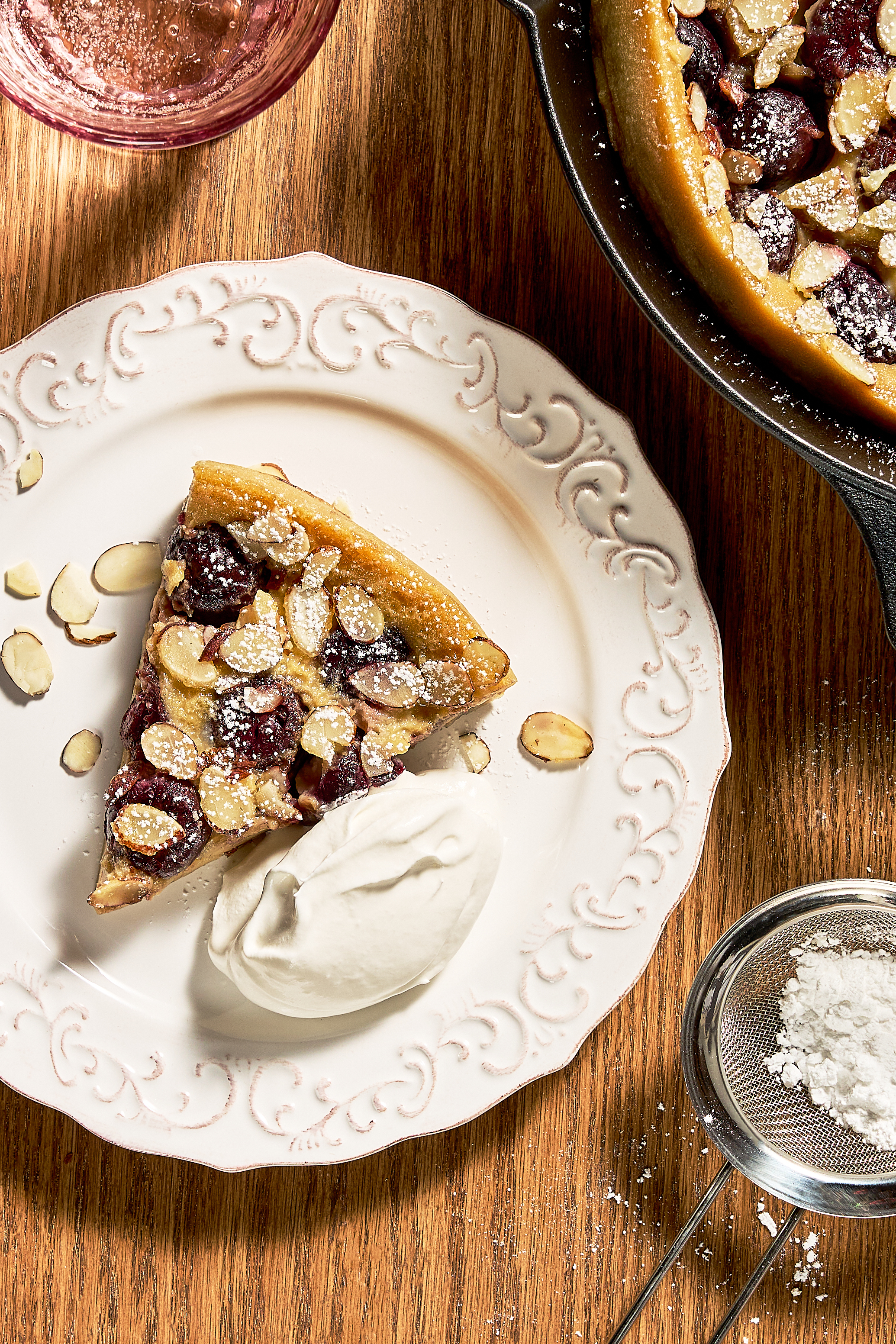 Clafoutis aux cerises entièrement cuit, garni d'amandes effilées, avec une pointe en moins et une deuxième coupée mais non retirée, présenté dans une poêle en fonte reposant sur une table en bois au grain prononcé.