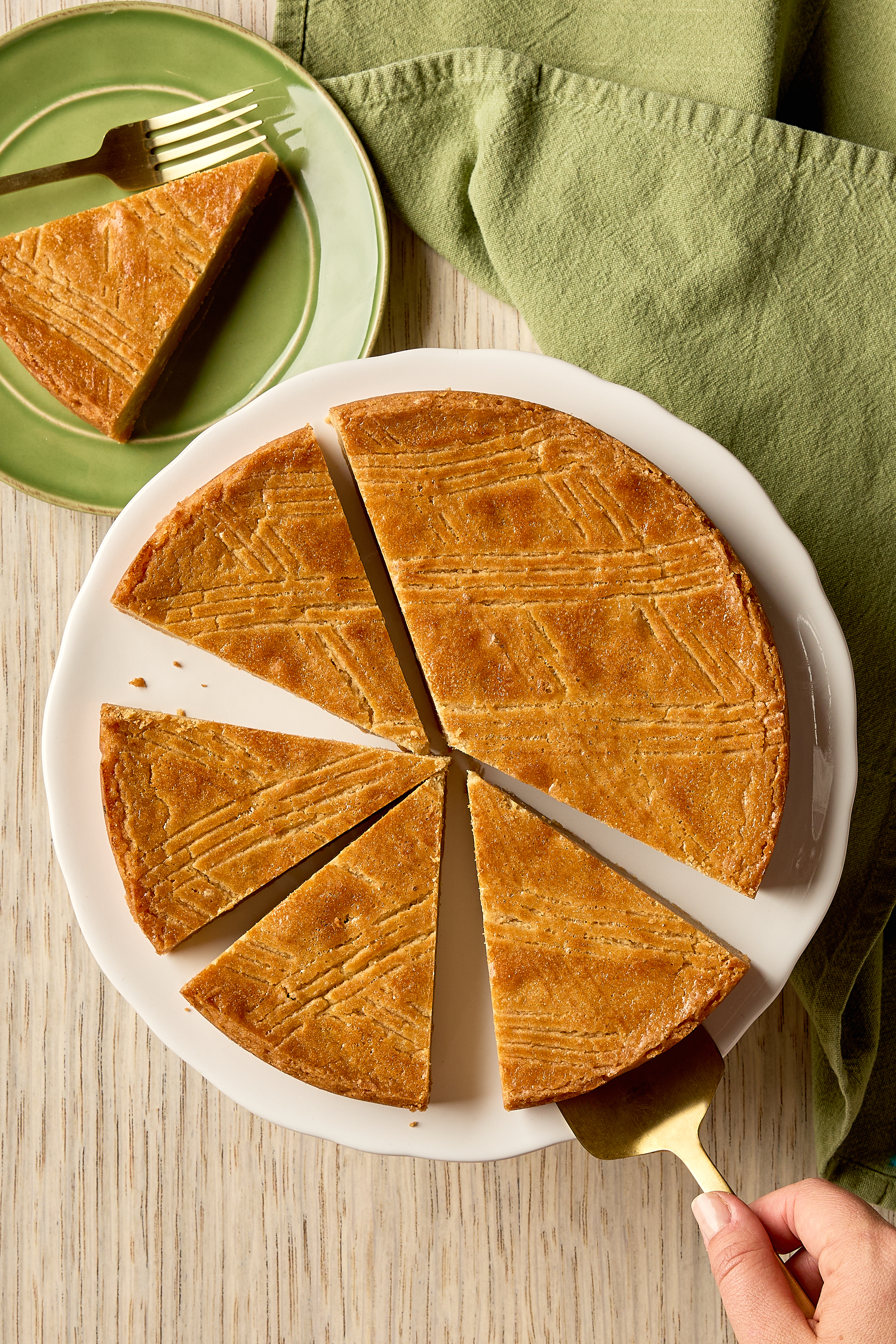 Overhead view of a sliced Brown Sugar Dutch Butter Cake on a white cake stand, set against a wooden background. A hand holds a gold cake server, lifting one slice.