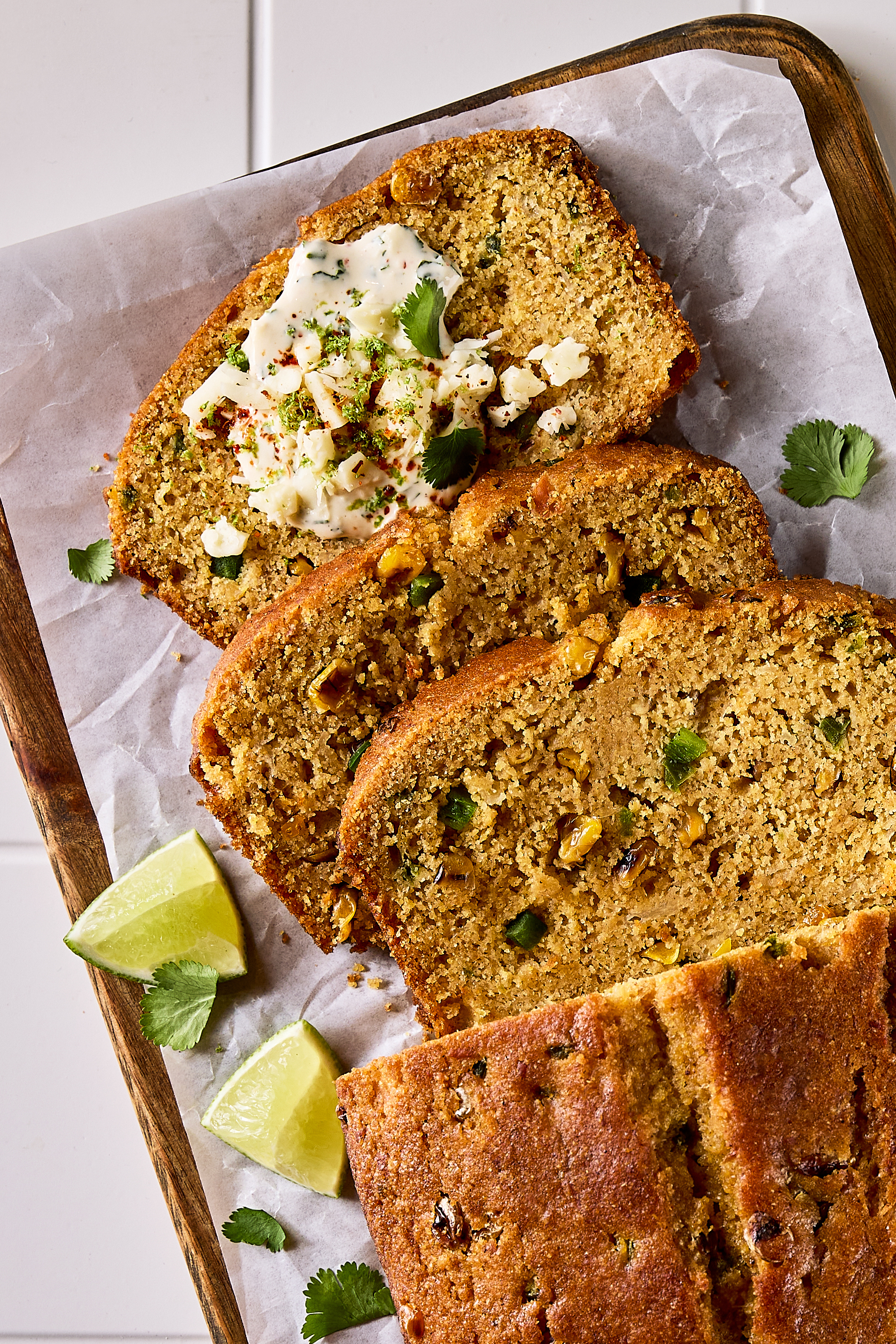 An elote cornbread loaf with three slices cut and fresh lime wedges on a cutting board, shown on a tiled kitchen counter