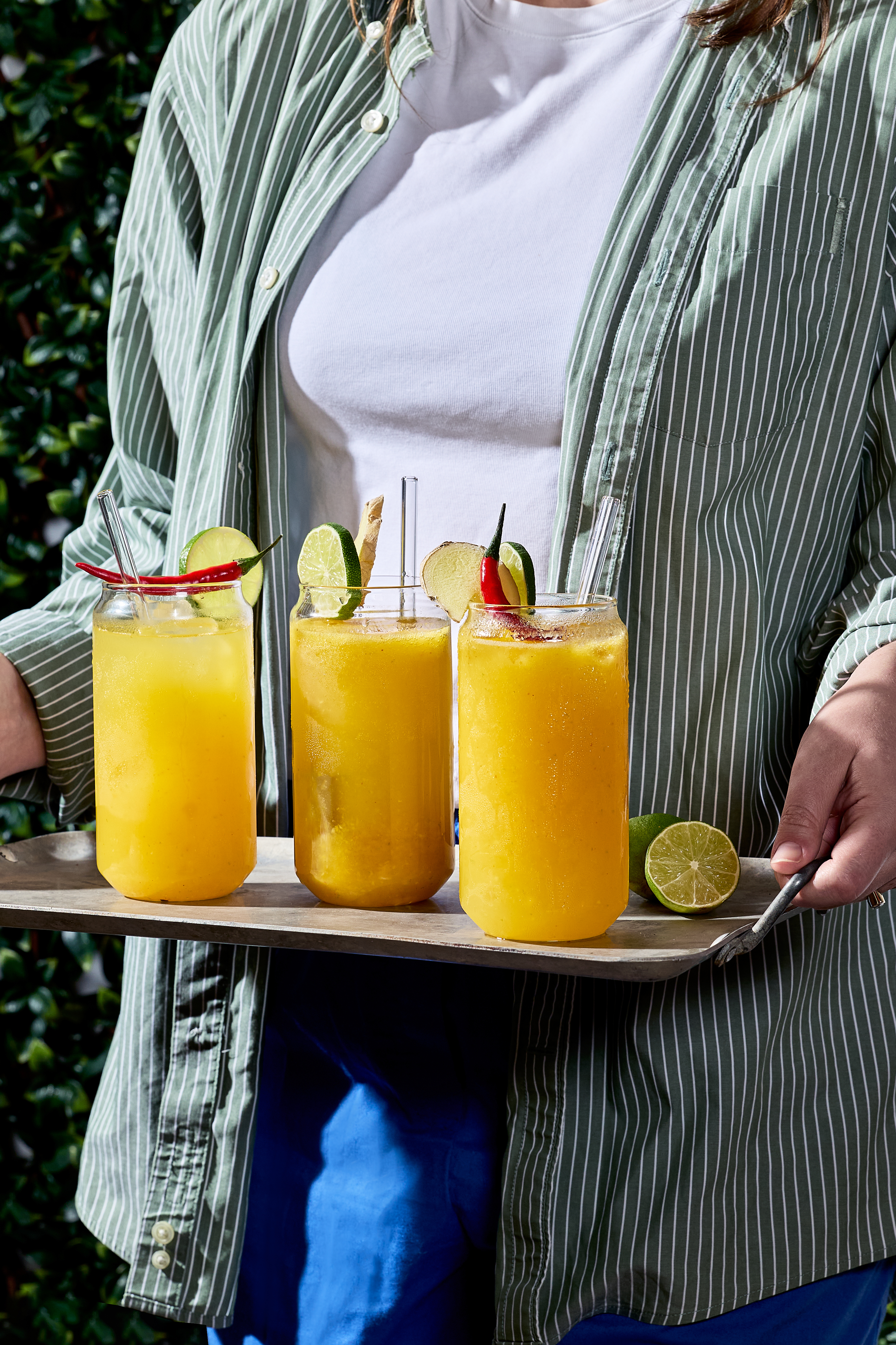 A woman with a striped shirt over a white t-shirt carries a tray outdoors of three glasses of sparkling mango ginger-chili refresher with glass straws, each garnished with a red chili pepper, a slice of ginger, and a slice of lime.