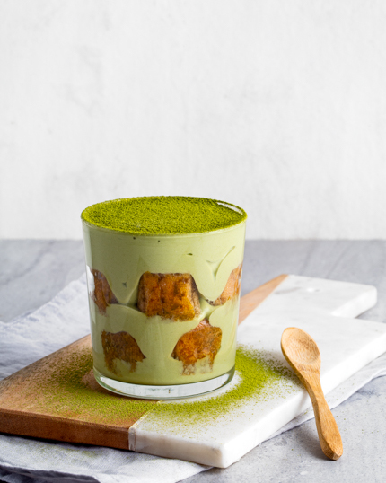 A glass cup of tiramisu sprinkled with green matcha powder shown with a wooden spoon