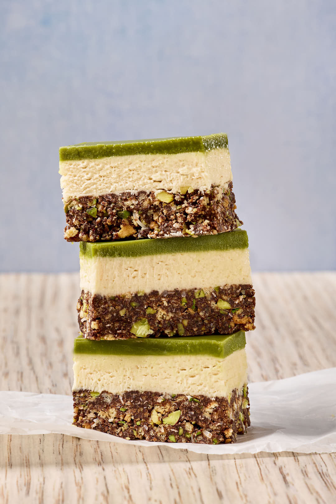 Three matcha Nanaimo bars stacked vertically, highlighting their layers of chocolate coconut base with pistachios, creamy vanilla custard filling, and a smooth green matcha topping, on a parchment-lined surface.