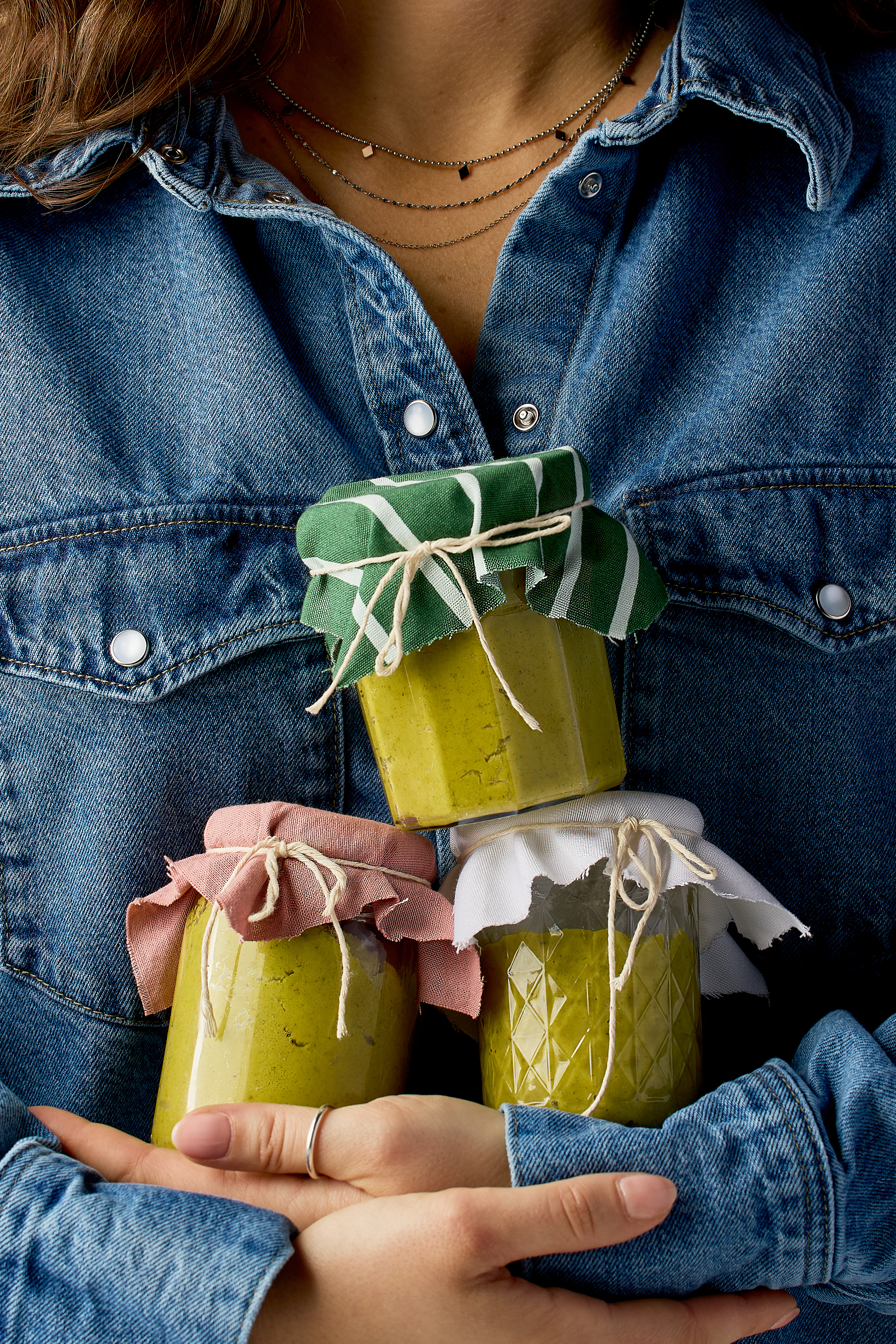 A woman in a denim shirt holding three jars of pistachio spread, each jar topped with cloth and tied off with string for gifting
