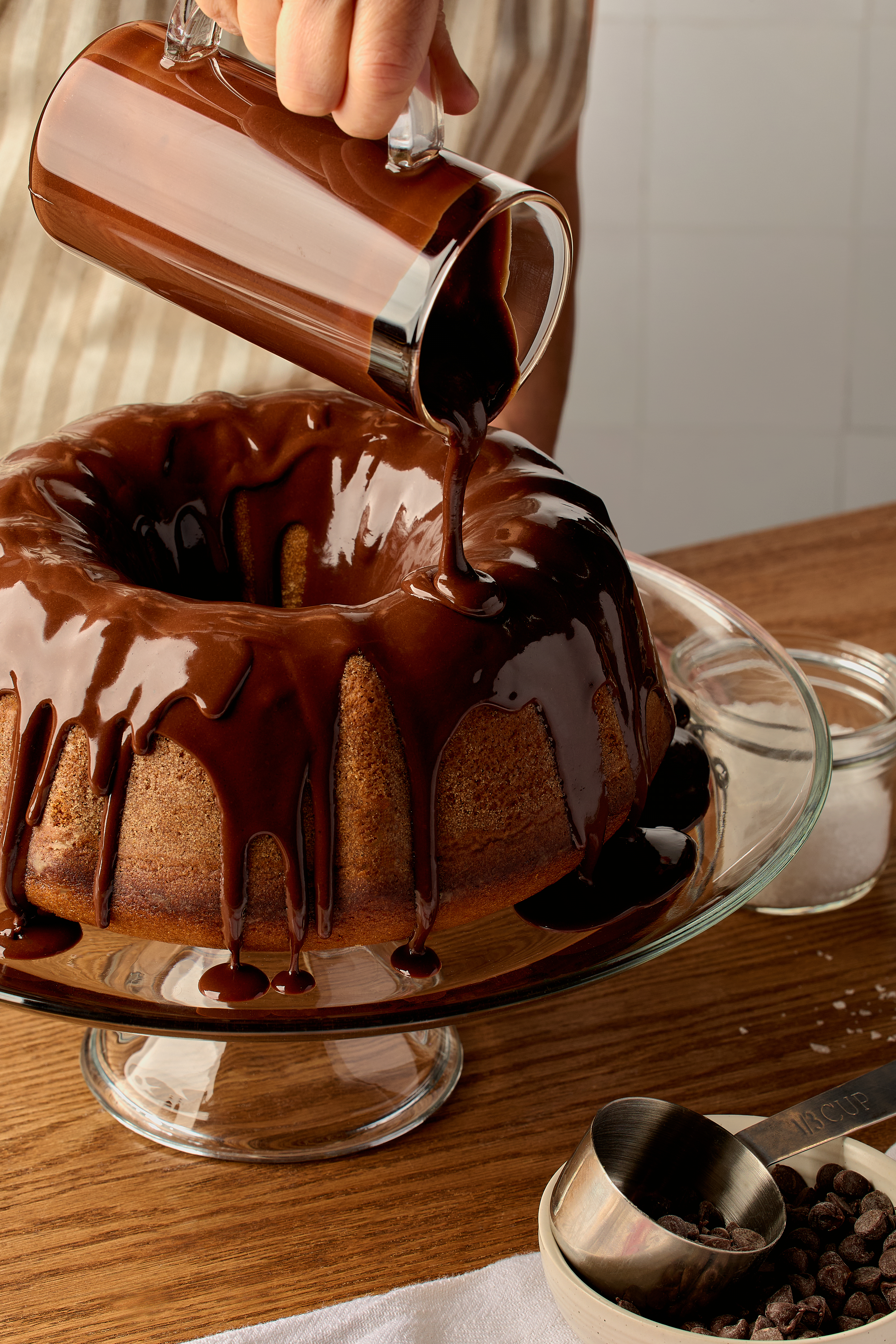 A Peanut Butter Bundt cake on a glass cake stand set on a wooden table, shown with a person pouring ganache over the cake from a glass pitcher