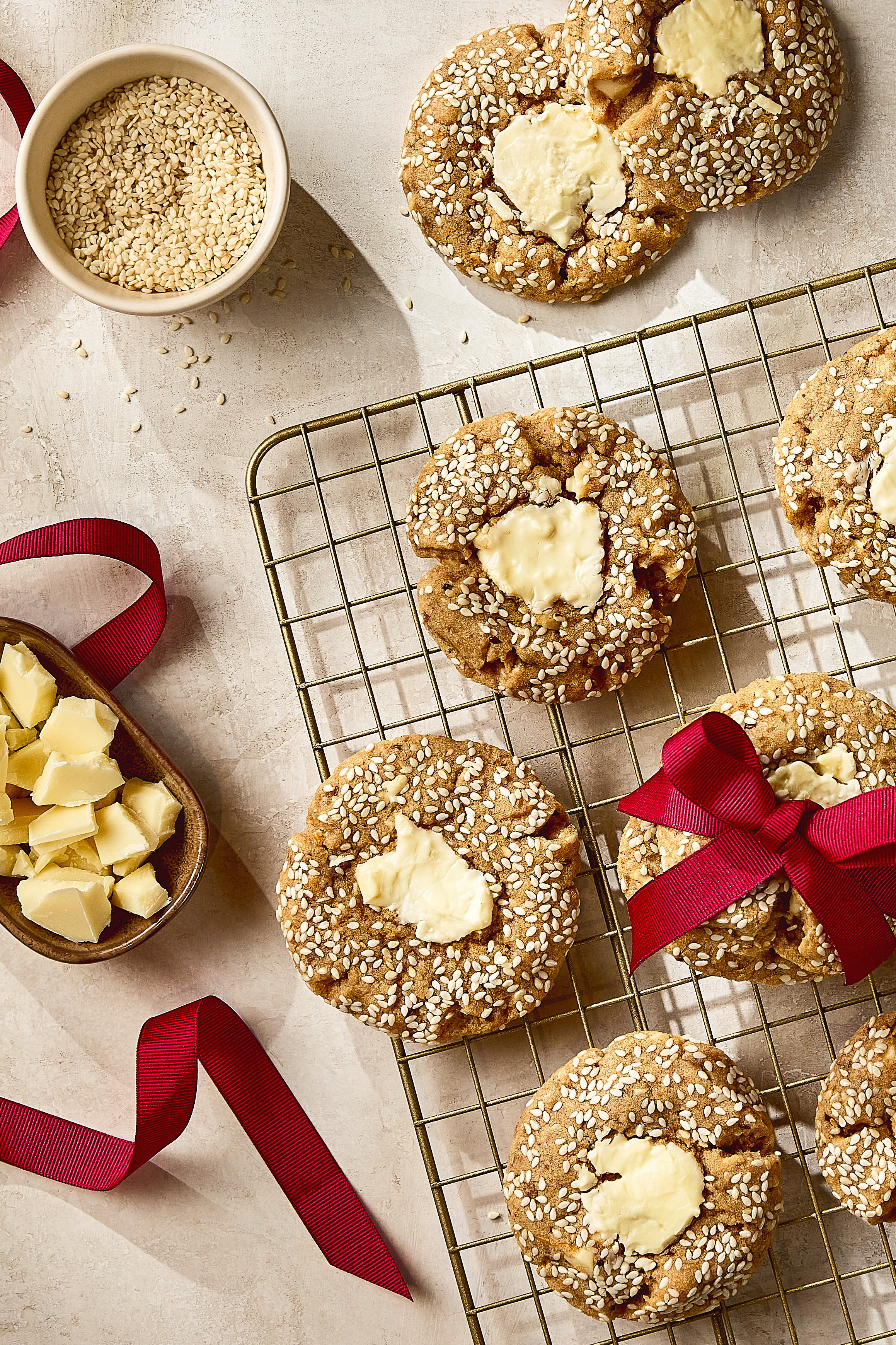 White chocolate chunk tahini cookies coated in sesame seeds cooling on a wire rack, with one cookie tied in a red ribbon. A bowl of sesame seeds, a dish of chopped white chocolate, and loose red ribbon are arranged around the cookies on a light textured surface.