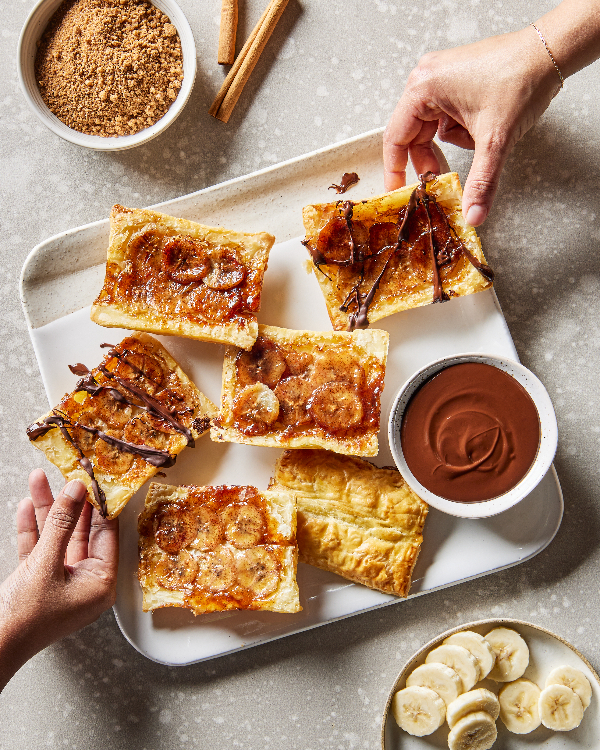 Four upside-down banana tarts, two on plates and two on a tray, shown with a bowl of brown sugar and cinnamon sticks.