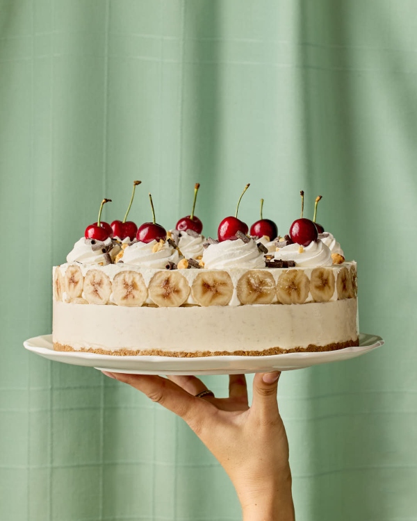 A hand holds up a banana split ice cream pie on a white plate in front of a soft mint green background. The pie features a graham cracker crust, a creamy frozen filling, a ring of banana slices around the sides, and is topped with swirls of whipped cream, maraschino cherries, chopped nuts, and chocolate shavings.