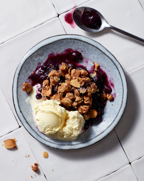 A bowl of blueberry crisp with vanilla ice cream shown with a spoon on a tile counter