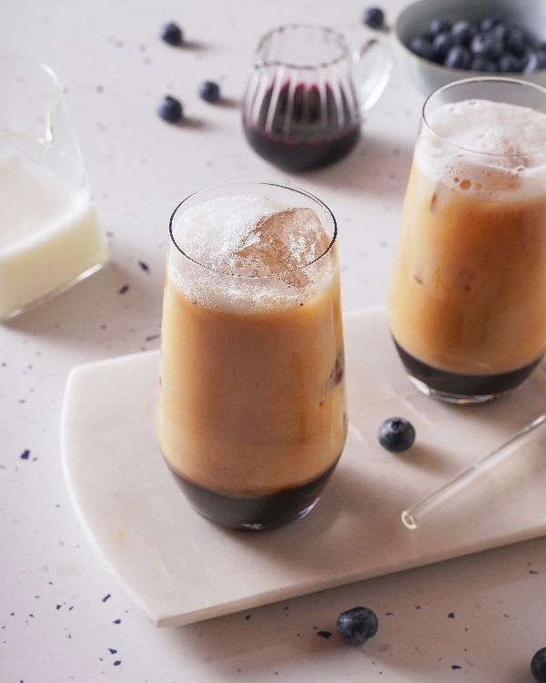 Iced coffee drinks served in tall glasses with visible layers of blueberry syrup, coffee, and milk. The drinks are topped with large ice cubes and placed on a white stone serving board. In the background, there is a small jug of milk, a glass pitcher of blueberry syrup, a bowl of fresh blueberries, and scattered blueberries on a speckled countertop.