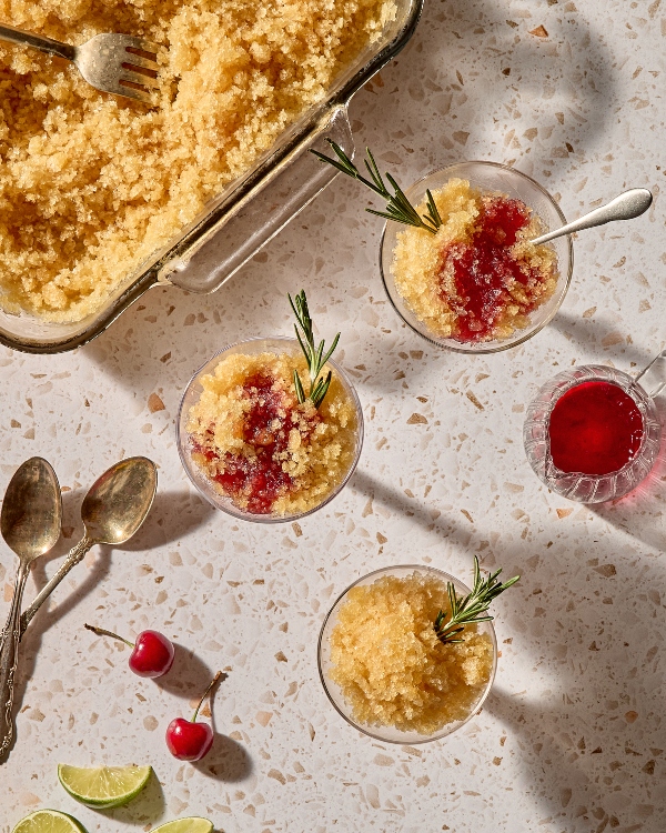Overhead view of three glass cups filled with a golden slushy dessert, some drizzled with a deep red syrup and garnished with rosemary sprigs. Nearby are vintage spoons, fresh cherries, lime wedges, and a glass pitcher of red cherry syrup. A large glass dish of the golden slush with a fork sits in the top left corner on a speckled terrazzo surface.