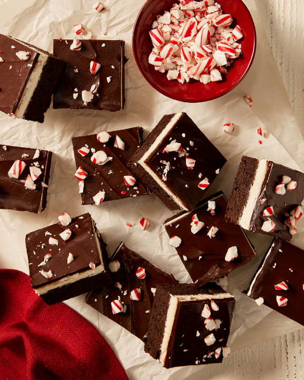 Chocolate mint brownies with a layer of mint cream and ganache top, each sprinkled with candy cane pieces, shown on wax paper on a white surface with a red napkin and a red bowl of candy cane pieces