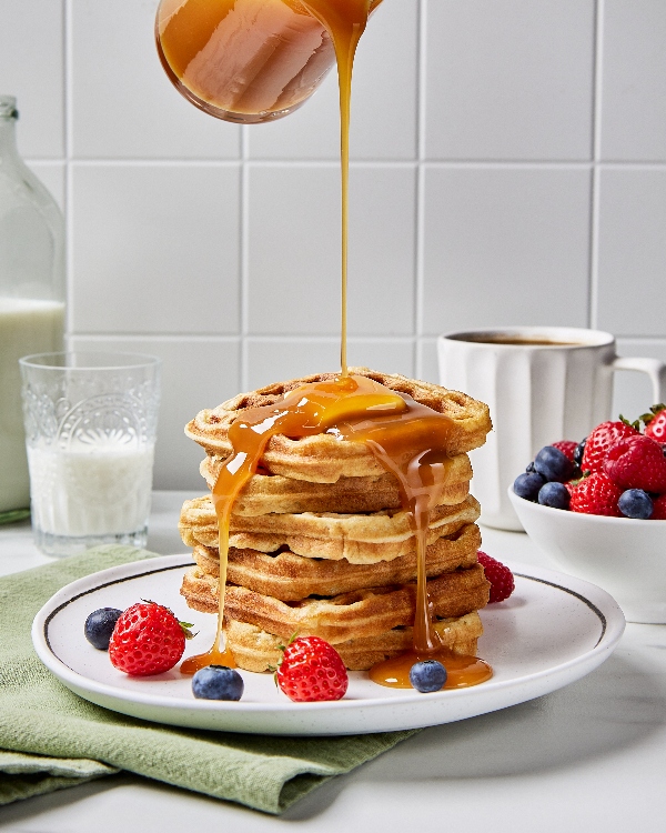 Syrup pouring from a height over a stack of buttermilk waffles on a white plate on a kitchen counter, shown served with a bowl of strawberries and blueberries, a cup of milk, and a mug of coffee.