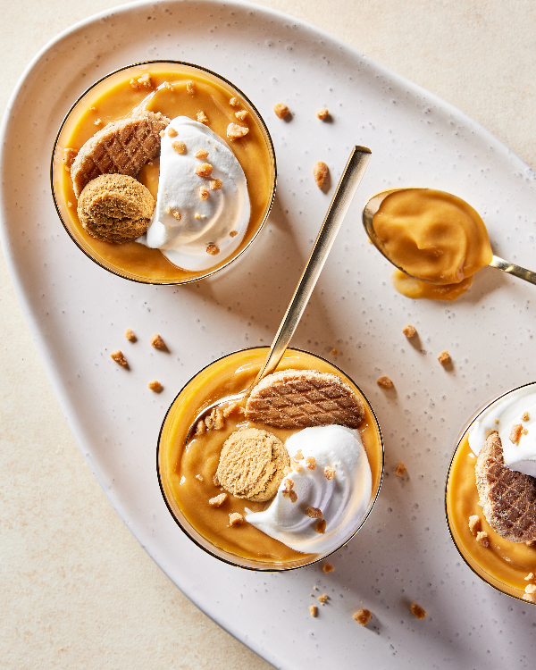 Top view of three glasses of butterscotch pudding on a small oval tray, each topped with whipped cream, toffee bits, and cookies.
