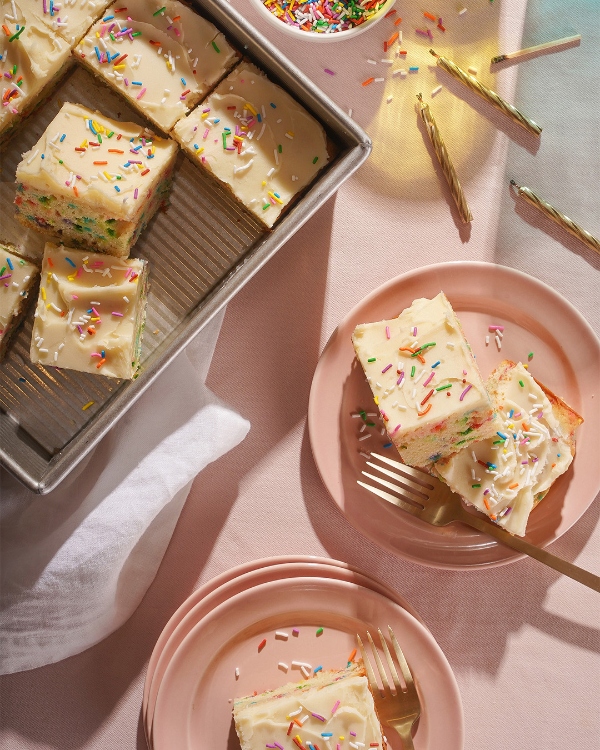 Frosted sheet cake cut into squares decorated with colorful sprinkles, some arranged in a baking pan and others plated on pink dishes with gold forks, along with scattered sprinkles and birthday candles on a light pink surface.