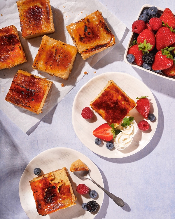 An overhead view of individual portions of crème brûlée with a caramelized sugar crust, some plain on parchment paper, and others garnished with fresh berries and whipped cream on small plates. A bowl of mixed berries sits in the upper right corner, and a spoon rests on one of the plated desserts.