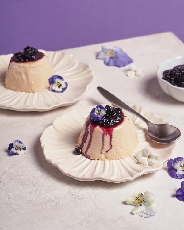 Two servings of Earl Grey panna cotta topped with blueberry compote and edible flowers, served on scalloped cream-colored plates with a spoon, against a soft purple and cream background.
