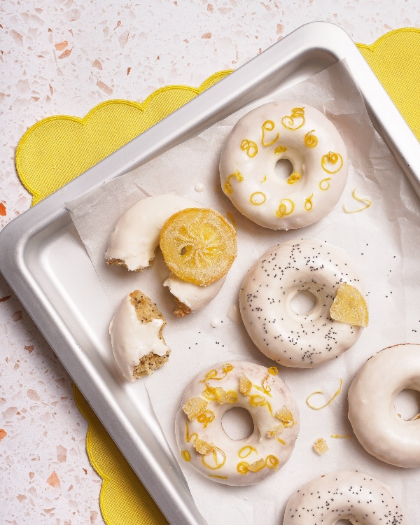 A baking sheet lined with parchment paper holding glazed lemon poppyseed doughnuts, garnished with lemon zest, candied lemon slices, and poppy seeds; one doughnut is partially eaten. The sheet sits on a terrazzo countertop with a yellow scalloped-edge placemat peeking out.