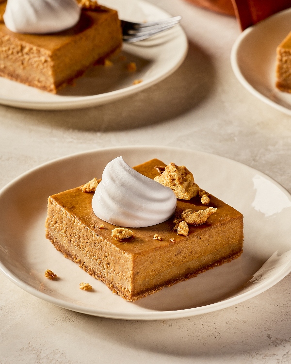 A single square of salted maple pumpkin dessert on a cream-colored plate, topped with a swirl of whipped cream and sprinkled with golden crumble pieces. Other plates with similar slices appear softly in the background, set on a neutral-toned table surface with warm, natural lighting.