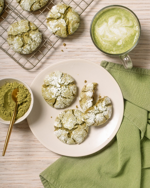 A top-down view of matcha crinkle cookies on a plate, some broken open, alongside a bowl of matcha powder with a gold spoon, a matcha latte in a glass mug, and more cookies on a wire rack, all on a light wood surface with a green napkin.