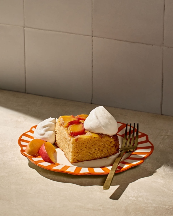 A single square of peach upside-down cake served on a red-and-white striped plate. The cake is topped with caramelized peach slices and dollops of whipped cream. Fresh peach wedges and a gold fork are placed beside the dessert on a beige surface with soft natural lighting.