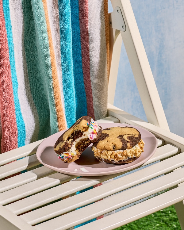 Two marbled chocolate and peanut butter ice cream sandwiches on a pale pink plate, resting on a white slatted lounge chair. One sandwich is rolled in rainbow sprinkles and bitten into, the other in chopped nuts. A colourful striped towel hangs over the chair back.