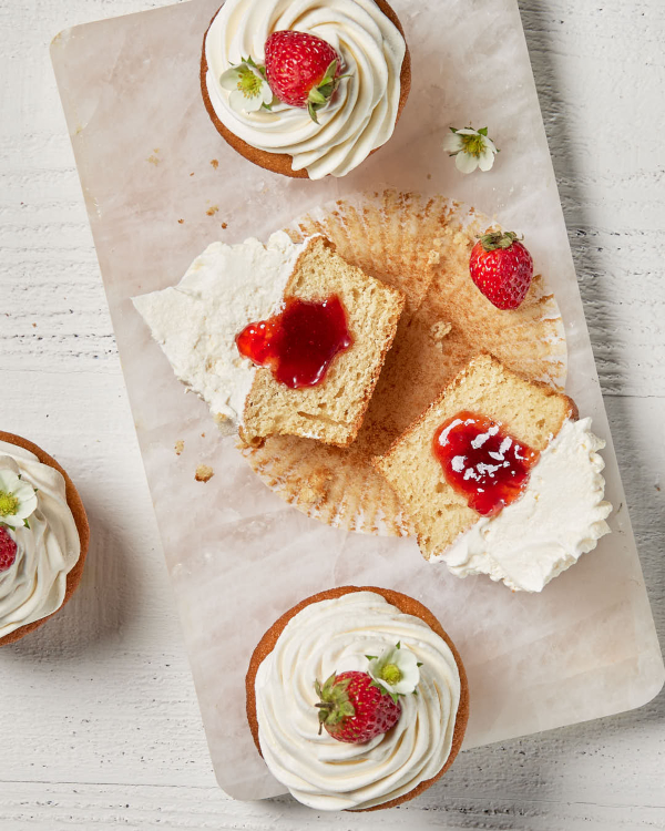 Four vanilla cupcakes topped with swirled icing and a whole strawberry, one shown sliced in half with strawberry sauce in the center on a granite cutting board.