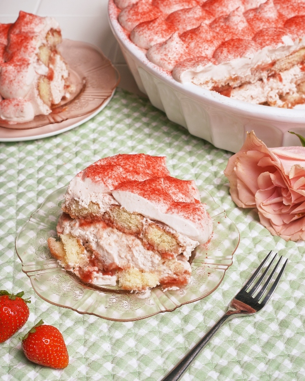 A slice of strawberries and cream tiramisu on an ornate glass plate, layered with sponge cake and a creamy filling and top. A second slice is in the background beside the rest of the dessert in a white baking dish, all sitting on a green gingham cloth with a fork, fresh strawberries, and pink roses.