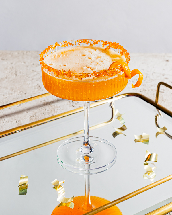 One glass of sparkling spiced ginger turmeric cocktail garnished with orange peel and rimmed with sugar on a mirrored serving tray