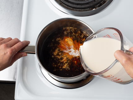 Pouring evaporated milk from a glass measuring cup into a pot of hot water and tea leaves