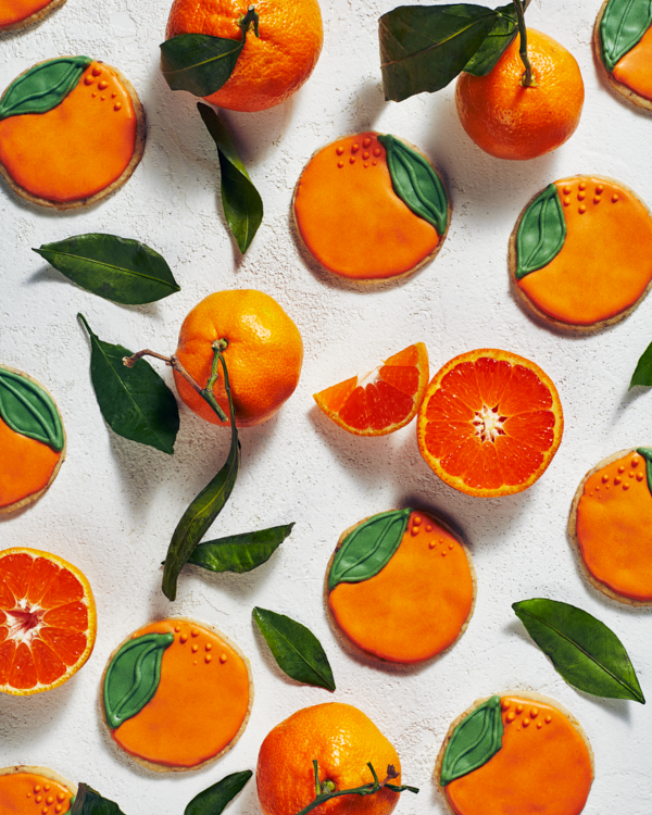 Shortbread cookies decorated to look like oranges shown with whole and sliced oranges and orange tree leaves.