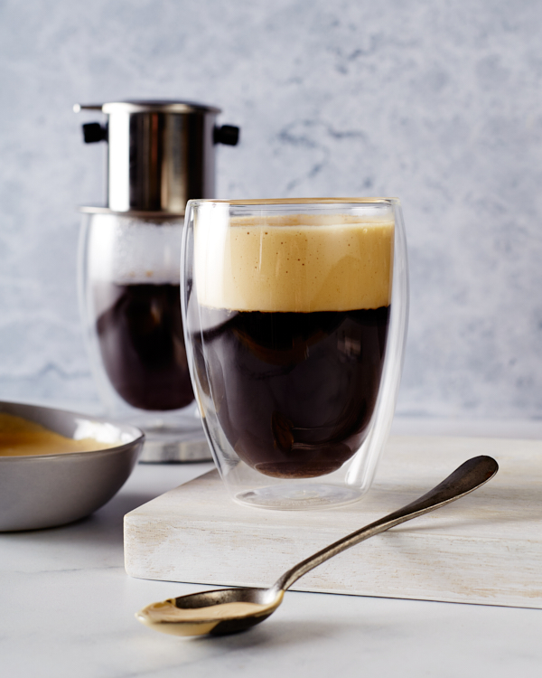 A glass of Vietnamese Egg Foam Coffee shown with a bowl of egg foam, a glass of coffee, and coffee-making equipment.