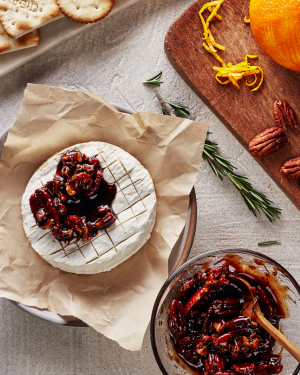 Baked brie topped with spiced candied pecans and Dark Brown sugar syrup and garnished with orange zest and rosemary, shown on a plate on a serving board with crackers, crostini, and sliced pears and apples, with wine glasses in the background.