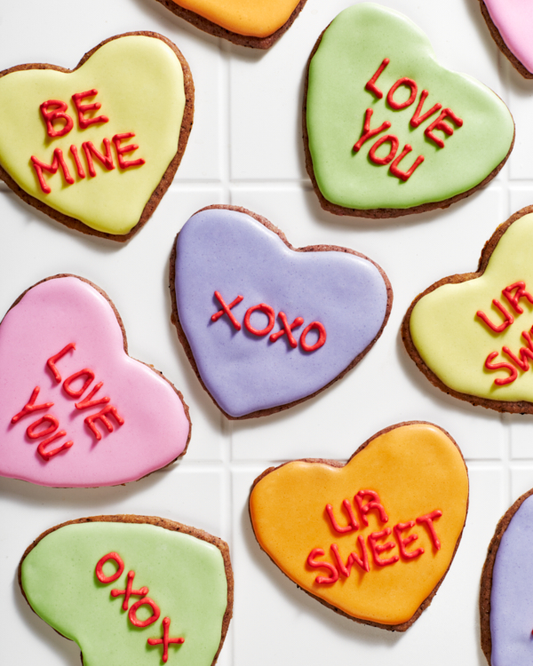 Heart-shaped sugar cookies decorated with royal icing in pastel coloulrs with romantic messages written in icing, shown on white tiles.
