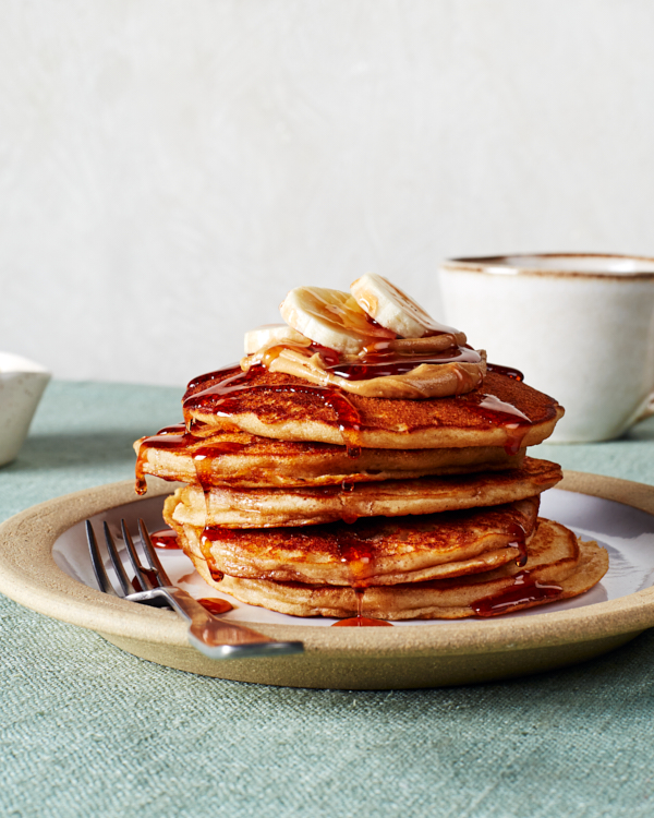 A stack of five peanut butter pancakes topped with syrup, sliced bananas, and peanut butter shown with a mug on a table