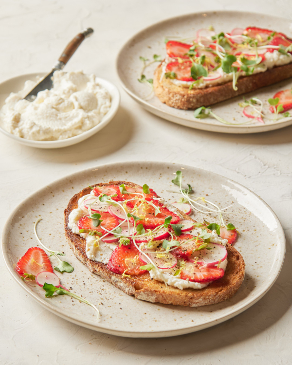 Two slices of toasted bread topped with whipped brown sugar ricotta, sliced strawberries and radishes, and microgreens, shown with a bowl of whipped brown sugar ricotta