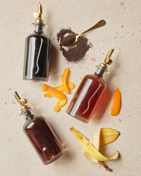 A bottle of coffee simple syrup, a bottle of orange simple syrup, and a bottle of banana simple syrup, shown with coffee grounds, orange peels, and banana peels 