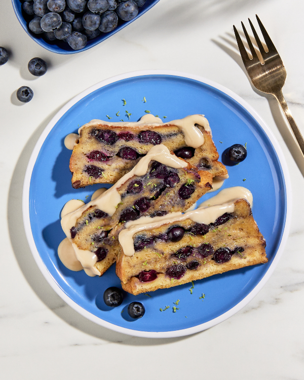 Two slices of loaf pan French toast with blueberries drizzled with maple glaze and shown with a cup of coffee, a dish of blueberries, and the partially sliced loaf 