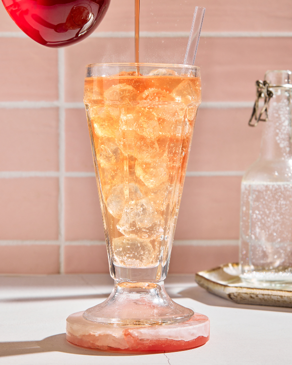 A parfait glass of old-fashioned cream soda on ice shown with a bottle of soda and pouring strawberry simple syrup from a pitcher into the glass 