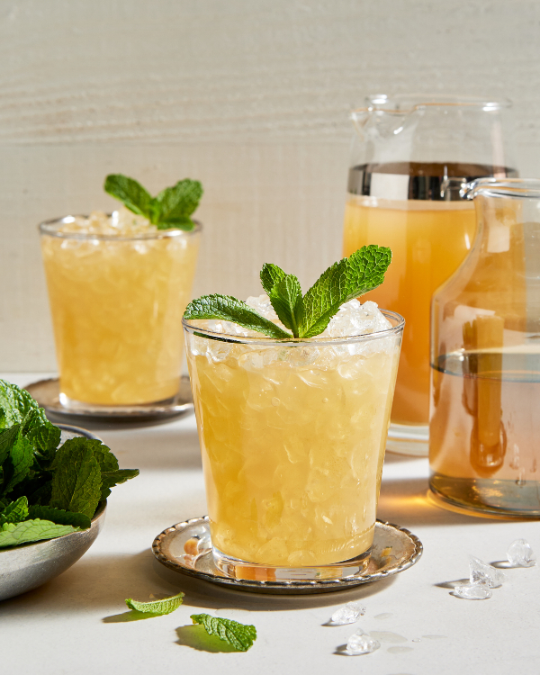 Two glasses of spirit-free mint julep with crushed ice and garnished with mint, shown with a bowl of mint leaves, a pitcher of prepared julep and a pitcher of mint simple syrup