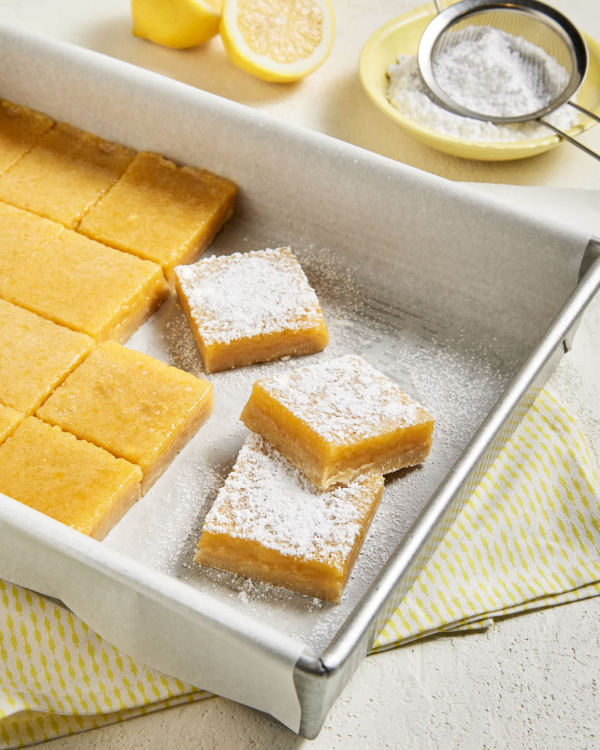 Cut lemon bars in a parchment-lined baking pan, three shown separated and dusted with icing sugar, with a cut lemon and a bowl of icing sugar with a sifter in the background.