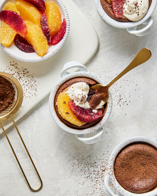 Overhead view of three personal-size molten chocolate lava cakes in white ramekins, two topped with whipped cream and cocoa powder and garnished with orange and blood orange segments, one plain, shown with a bowl of orange and blood orange segments and a sifter of cocoa powder.