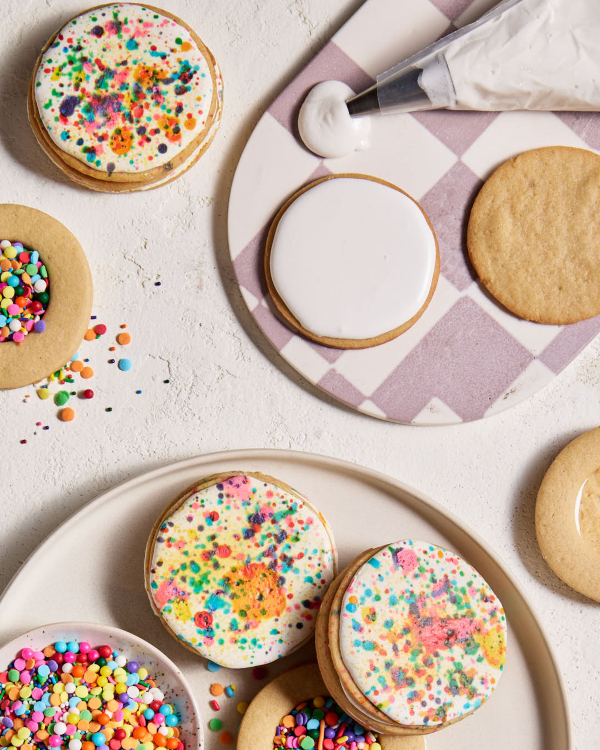 Three iced pinata sugar cookies, shown with two incomplete cookies filled with candy confetti and sprinkles, a bowl of candy confetti and sprinkles, and a piping bag filled with and leaking white icing on a white and mauve checkered tray.