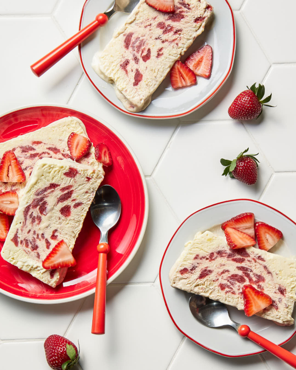 Slices of strawberry-elderflower semifreddo on three plates, two white with red trim and one red with white trim, each with a red spoon, and shown with whole strawberries scattered on a white tile counter.