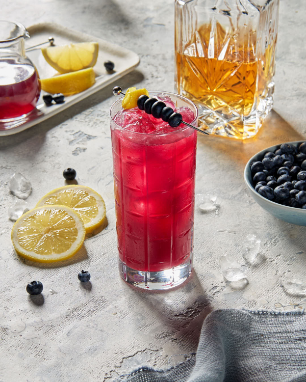 A highball glass of blueberry whisky fizz on ice garnished with a skewer of blueberries, shown with a decanter of whisky, a pitcher of simple syrup, lemon wedges and wheels, and a bowl of blueberries.