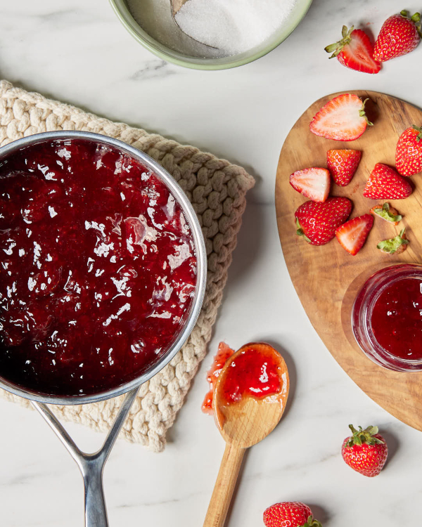 A pot of fresh strawberry jam on a dish cloth, shown with sliced strawberries, a wooden spoon, and a bowl of granulated sugar.