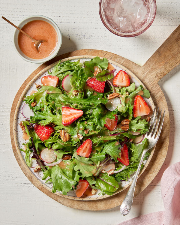 A green leaf salad on a plate on a wooden serving board, garnished with sliced strawberries, shown with a bowl of strawberry vinaigrette.