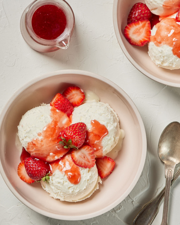 Two pale pink bowls of vanilla ice cream topped with sliced strawberries and strawberry sauce.